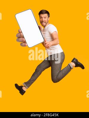 Aufgeregt Mann springen, zeigen Finger auf die Kamera, zeigt Smartphone mit leerem Bildschirm über orangenen Hintergrund, Mockup Stockfoto