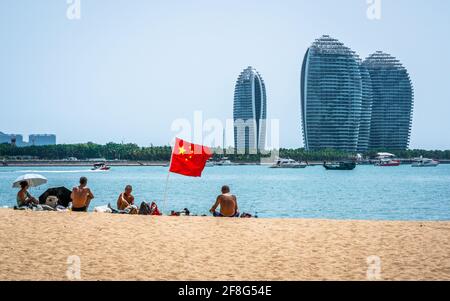 Sanya China , 24. März 2021 : Gruppe von Menschen um eine chinesische Flagge am Strand von Sanya und Blick auf Gebäude der Phoenix-Insel in der Bucht von Sanya Hainan Stockfoto
