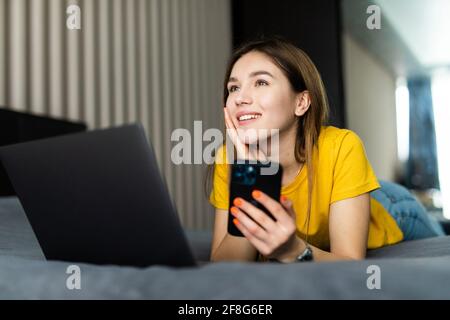 Junge Frau, die in einem weißen Hemd auf dem Bett sitzt und eine Tasse Kaffee in der Hand hält. Schöne Frau nutzt Laptop, um Online-Inhalte und Online-Schulungen anzusehen. Stockfoto