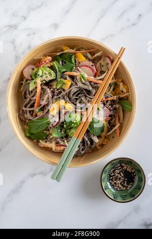 Flatlay eines veganen Sesam Soba Nudelsalats mit viel Gemüse mit Essstäbchen auf weißem Marmorboden, glutenfreies Rezept Stockfoto