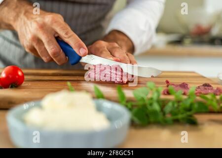 Eine Nahaufnahme der Hände des Küchenchefs, die während der Vorbereitungsphase Carpaccio-Fleisch auf einem Holzbrett in dünne Stücke schneiden. Verschiedene kleine Küche b Stockfoto