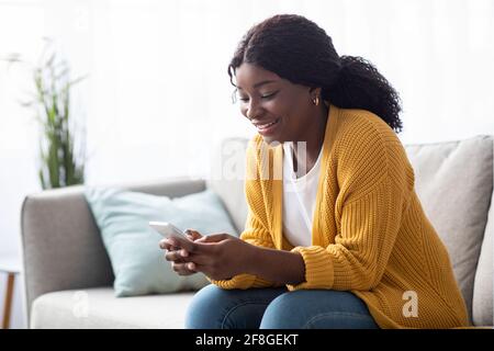 Lächelnde schwarze Dame, die auf der Couch sitzt, mit dem Smartphone Stockfoto