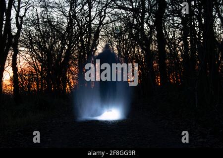 Ein Horrorkonzept einer verschwommenen gruseligen Figur, umgeben von Lichtern. Steht in einem dunklen Winterwald kurz nach Sonnenuntergang. Stockfoto