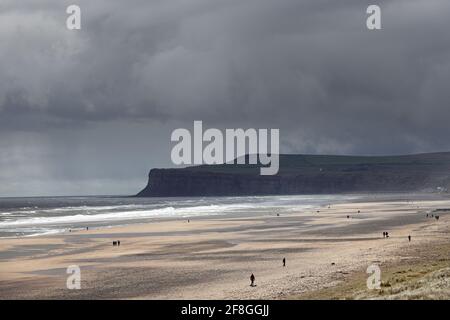 Menschen, die am Strand spazieren, während Sturmwolken über Saltburn-by-the-Sea und Huntcliff, North Yorkshire, Großbritannien, Rollen Stockfoto