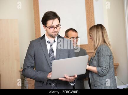 Happy serious jungen Geschäftsmann mit einem modernen Laptop-Computer Mit seinem Team im Hintergrund Stockfoto