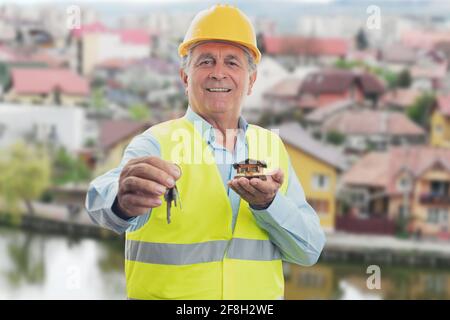 Fröhlicher, alter Mann, der in gelben Hartmützen zuhause ist Schlüssel und Miniatur-Holzhaus mit Gebäuden Hintergrund Stockfoto