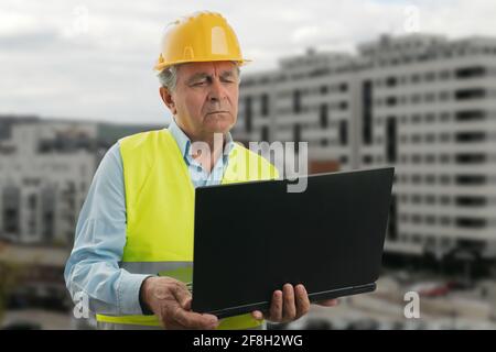Älterer Baumeister mit gelbem Schutzhelm und fluoreszierender Weste Mit konzentrierten Blick auf Notebook-pc-Builder auf das Bauen Hintergrund Stockfoto