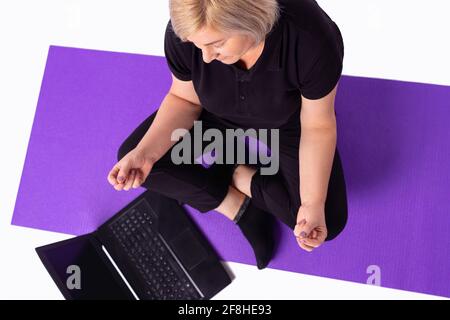 Ältere gesunde ruhige Frau blonde Haare in Sportswear meditieren Online-Kurs auf Laptop zu Hause beobachten. Fit sportlich ältere Menschen tun Yoga-Training Fitness Stockfoto