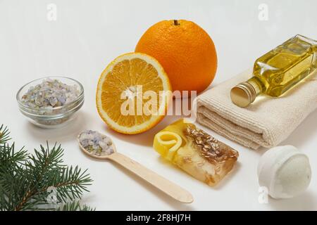 Schneiden Sie Orange mit einem ganzen Stück, einem Frottee-Handtuch, einer Flasche mit Aromatherapieöl, hausgemachter Seife, einer Badebombe und einem Holzlöffel mit Meersalz auf dem weißen Bac Stockfoto