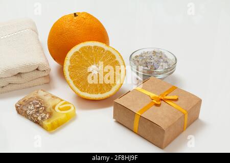 Schneiden Sie Orange mit einem ganzen Stück, einem Frottee-Handtuch, hausgemachter Seife, einer Geschenkbox und einer Glasschale mit Meersalz auf dem weißen Hintergrund. Wellness-Produkte und Zubehör Stockfoto