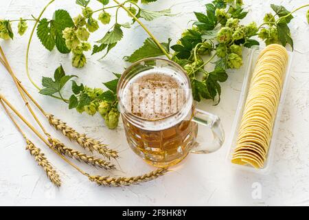 Glas Becher Bier mit Ohren von Gerste, Kartoffelchips und Hopfenzweige auf dem weißen strukturierten Hintergrund. Draufsicht. Stockfoto