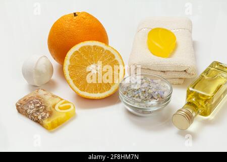 Schneiden Sie Orange mit einem ganzen Stück, einem Frottee-Handtuch, einer Flasche mit Aromatherapieöl, hausgemachter Seife, einer Badebombe und einer Glasschale mit Meersalz auf dem weißen Bac Stockfoto
