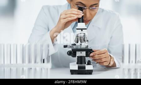 Fokussierter afroamerikanischer Wissenschaftler, der in der Nähe von Reagenzgläsern durch das Mikroskop schaut Im Labor Stockfoto
