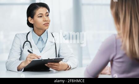 afroamerikanischer Arzt im weißen Mantel schriftlich Diagnose in der Nähe des Patienten Im unscharfen Vordergrund Stockfoto