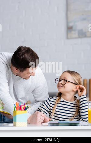Lächelndes Mädchen in einer Brille, das den Vater ansieht, während er Hausaufgaben macht, verschwommener Vordergrund