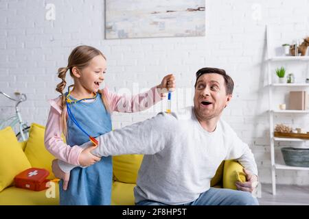 Aufgeregt Mädchen macht Injektion mit Spielzeug Spritze zu Vater vortäuscht Erschrocken Stockfoto