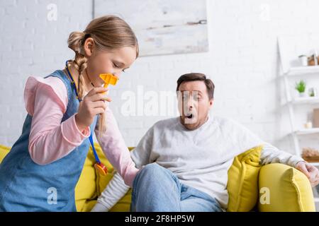Mädchen hält Spielzeug Reflex Hammer in der Nähe Knie des Vaters sitzt auf der Couch mit offenem Mund, verschwommener Hintergrund Stockfoto