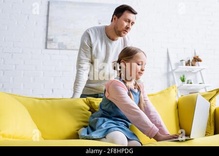 Überraschender Mann, der die Tochter auf dem Sofa beim Tippen auf dem Laptop ansah Stockfoto