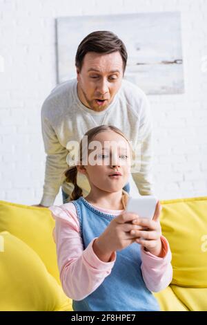 Staunender Vater in der Nähe der Tochter, die zu Hause auf dem Smartphone chattet Stockfoto