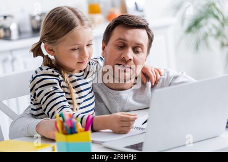 Vater hilft Tochter Hausaufgaben in der Nähe von Laptop auf verschwommenem Vordergrund Stockfoto