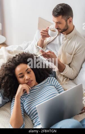 Blick aus der Perspektive einer afroamerikanischen Frau, die auf dem Bett liegt Und mit Laptop in der Nähe von Freund mit Smartphone trinken Kaffee Stockfoto