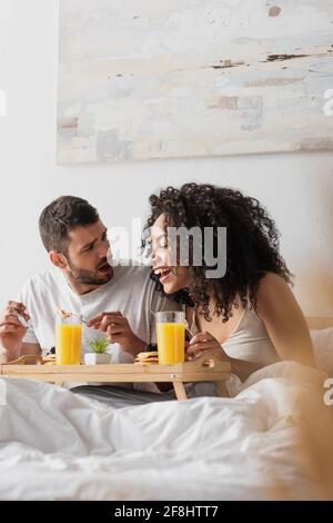 Schockierter bärtiger Mann, der eine glückliche afroamerikanische Freundin ansieht Frühstück Stockfoto