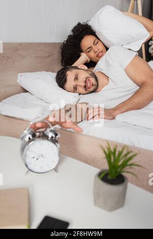 Unzufriedener Mann, der den Wecker in der Nähe einer afroamerikanischen Freundin erreichte Bett Stockfoto