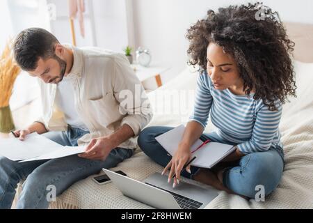 afroamerikanische Frau mit Notebook mit Laptop in der Nähe von bärtigen Freund Aufbewahrung von Papieren Stockfoto