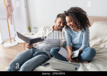 Glückliche afroamerikanische Frau mit Notebook mit Laptop in der Nähe von bärtig Freund hält Ordner im Schlafzimmer Stockfoto