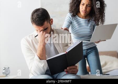 Mann mit Notizbuch und Deckblatt in der Nähe besorgt afroamerikanischen Freundin auf unscharfem Hintergrund Stockfoto