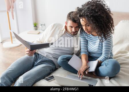 afroamerikanische Frau mit Notebook mit Laptop in der Nähe von bärtigen Freund Ordner im Schlafzimmer halten Stockfoto