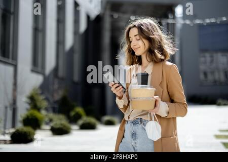Frau mit Schutzmaske auf der Hand, die allein in der leeren Straße in der Nähe des Bürogebäudes mit Essen zum Mitnehmen läuft. Über Telefon. Stockfoto