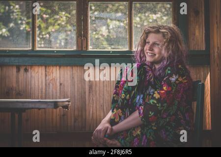 Liebenswert lächelnd glückliches Mädchen mit langen blonden Haaren sitzend auf Stuhl hinter dem Fenster und entspannend Stockfoto