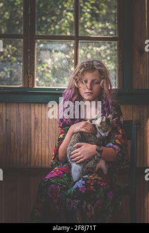 Entzückendes Kind mit langen blonden Haaren, die auf dem Stuhl hinter sich sitzen Das Fenster und entspannen, während man eine Katze umarmt Stockfoto