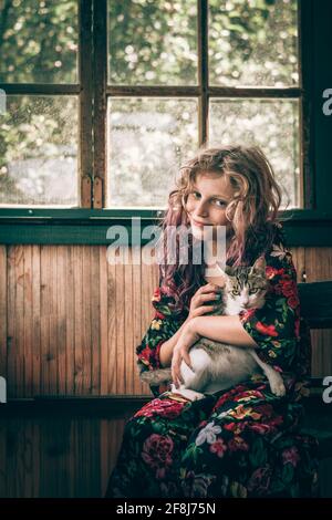 Entzückendes Kind mit langen blonden Haaren, die auf dem Stuhl hinter sich sitzen Das Fenster und entspannen, während man eine Katze umarmt Stockfoto