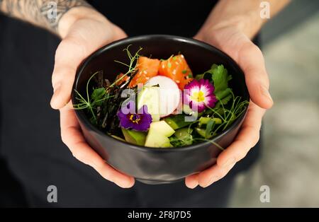 Ein Mann hält eine Schüssel Poke-Salat mit Lachs, Avocado, Gurke, Reis, Tamarindensauce, Mangosauce, Nori, Limette, Sesam, Koriander. Asiatischer Seafood-Salat Stockfoto
