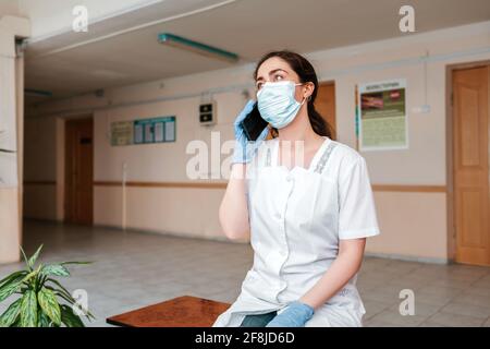 Coronavirus.EIN Arzt in einem Laborkittel, einer medizinischen Maske und Handschuhen, der im Flur sitzt und am Telefon anruft. Das Konzept der Heldenärzte Stockfoto