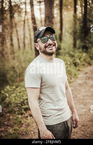 Mann mit Mütze und Sonnenbrille lächelt in einem wunderschönen Wald Stockfoto