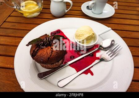 Schokoladenfondant mit Vanilleeis und Rotwaldfrucht-Sauce auf Teller mit Besteck, Kaffee, Milch und Glas Wasser auf Holztisch. Stockfoto