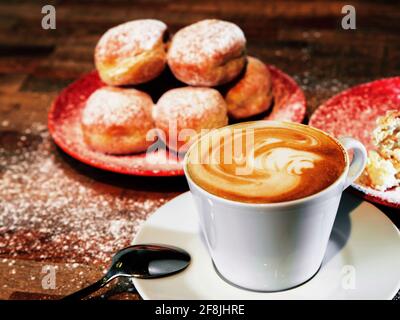 Tasse flachen weißen (Espresso mit Milchschaum) Kaffee, Gruppe von gerundeten frittierten Kuchen (Donuts) aus Teig mit Marmelade auf roten Tellern auf Holztisch gefüllt. Stockfoto