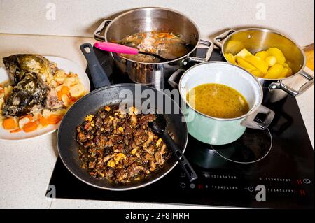 Zubereitung der Mahlzeit in vielen Töpfen auf Keramikherd in der Küche. Kochende Fischsuppe mit Gemüse, Kartoffeln, Karpfenkopf, Körper und Innereien. Stockfoto