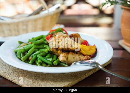 Gebratenes Schweinefilet, eingewickelt in Speck mit grünen Bohnen und Paprika auf weißem Teller mit Gabel, Korb mit Besteck auf Holztisch, im Freien. Stockfoto