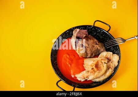 Gekochtes Rindfleisch-Shin, würzige Tomatensoße und Knödel auf ländlicher schwarzer Pfanne mit Gabel auf gelbem Hintergrund, Nahaufnahme. Diese Mahlzeit ist in Tschechien am beliebtesten. Stockfoto
