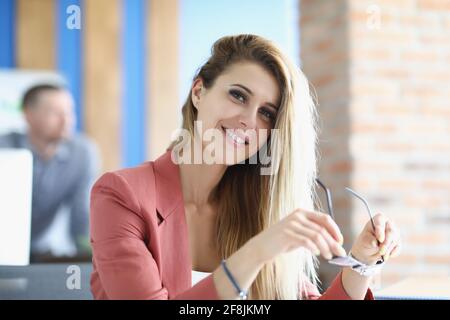 Junge Frau im Anzug sitzt am Tisch und hält eine Brille Für Vision in ihren Händen Stockfoto