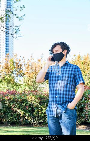 Porträt eines jungen hispanischen Mannes mit einer Stoffmaske, der während der Coronavirus-Pandemie, dem Covid, einen Anruf mit einem Smartphone in einem Stadtpark abgab Stockfoto