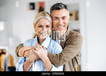 Nahaufnahme eines hübschen kaukasischen Ehepaares, glücklichen Mannes und Ehefrau in lässiger, stilvoller Kleidung, zu Hause stehend, umarmend, lächelnd direkt in die Kamera schauend Stockfoto