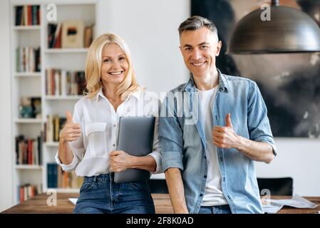 Porträt von zwei erfolgreichen Geschäftskollegen. Eine elegante Geschäftsfrau und ein selbstbewusster Geschäftsmann stehen in einem modernen Büro neben einem Schreibtisch, schauen auf die Kamera, zeigen Daumen nach oben, lächeln Stockfoto