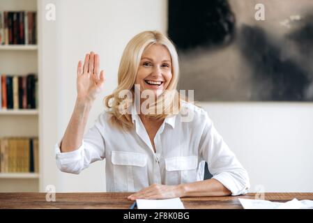 Virtuelle Online-Meetings. Fröhliche schöne kaukasische Erwachsene blonde Frau im Gespräch auf einer Videokonferenz, Begrüßung der Gesprächspartner mit einer Handbewegung, lächelnd freundlich, Blick auf die Webcam Stockfoto
