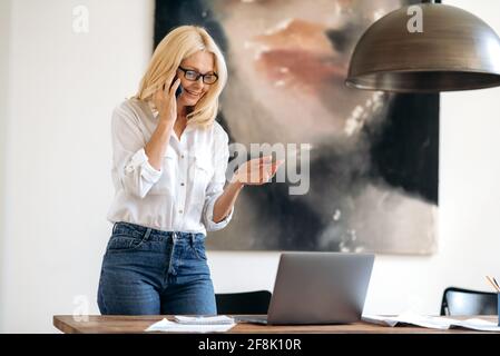 Erfolgreiche, elegante, vielbeschäftigte kaukasische Geschäftsfrau am Arbeitsplatz, stilvoll gekleidete, Erwachsene blonde Dame, die mit einem Kollegen auf dem Smartphone spricht, steht neben dem Schreibtisch, blickt auf den Laptop und lächelt freundlich Stockfoto
