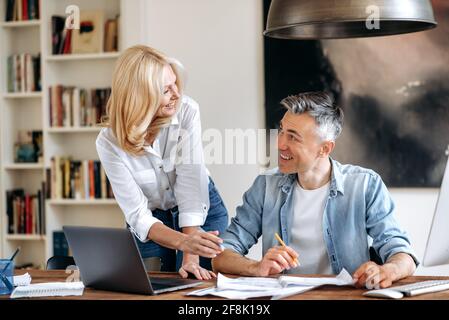 Zwei erfolgreiche kaukasische Geschäftskollegen mittleren Alters. Eine elegante Geschäftsfrau und ein selbstbewusster Geschäftsmann sitzen am Arbeitsplatz, arbeiten zusammen, diskutieren Arbeitsfragen, kommunizieren, lächeln Stockfoto